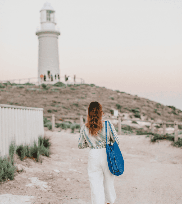 A Woman Is Walking On The Beach Near A Lighthouse.