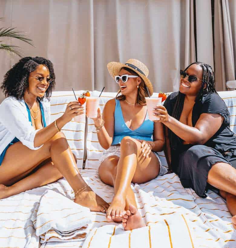 Three women sitting on a bed with drinks.