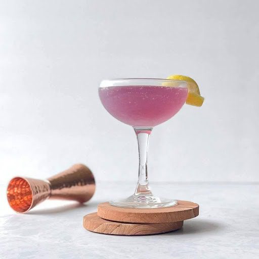 A pink cocktail in a glass with a lemon slice on top.
